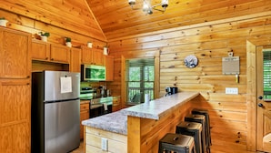 A wooden cabin kitchen with a stainless steel refrigerator, wooden cabinets, a granite countertop, bar stools, and a clock on the wall. - Great Cabin sleeps 8, New Furnishings, Loft w/ Arcade Games, Hot Tub & Fireplace (Sevierville)