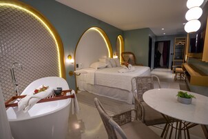 A modern hotel room with a large bed, a sitting area with a table and chairs, a bathtub, and a vanity with a mirror.