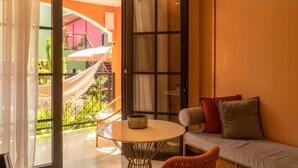 A room with a sliding glass door leading to a balcony with a hammock and plants. There is a round table with a bowl on it and a sofa with cushions.