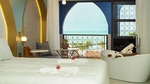 A hotel room with a large bed, a sitting area with a view of the ocean, a bathtub, and a balcony with a railing.