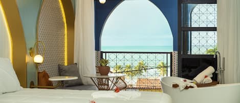 A hotel room with a large bed, a sitting area with a view of the ocean, a bathtub, and a balcony with a railing.