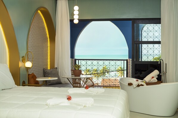 A hotel room with a large bed, a sitting area with a view of the ocean, a bathtub, and a balcony with a railing. - Exotica Boutique Hotel (Tibau do Sul)