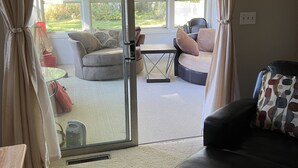 A living room with a glass door leading to a patio area. There is a black leather sofa, a small table with a red bag, and a grey sofa with cushions. - Park-like and sports bar - close to MSU (Lansing)