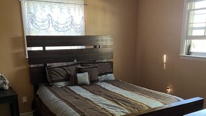 A bedroom with a wooden bed, a nightstand, a window with curtains, and a wall-mounted light fixture. - Park-like and sports bar - close to MSU (Lansing)