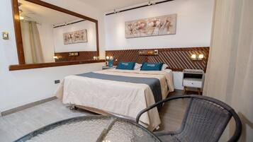 Una habitación de hotel con una cama grande, una mesita de noche, una silla, una mesita de vidrio, un espejo y un cuadro floral en la pared.