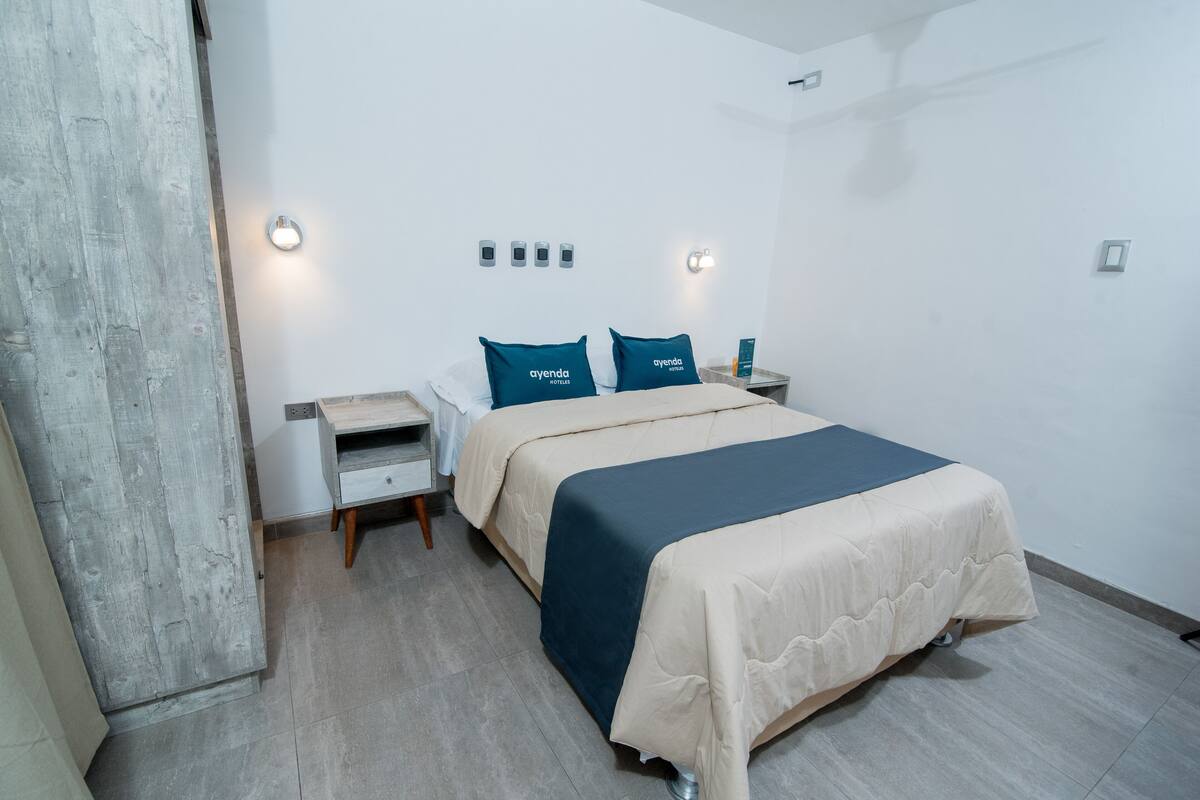 A hotel room with a bed, two pillows with the hotel name, bedside tables, and a wall-mounted light fixture.