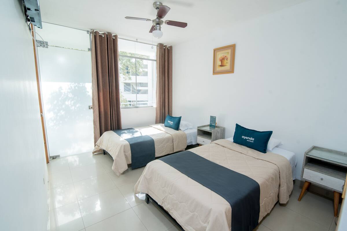 A hotel room with two beds, a ceiling fan, a nightstand, a small table, and a window with curtains.
