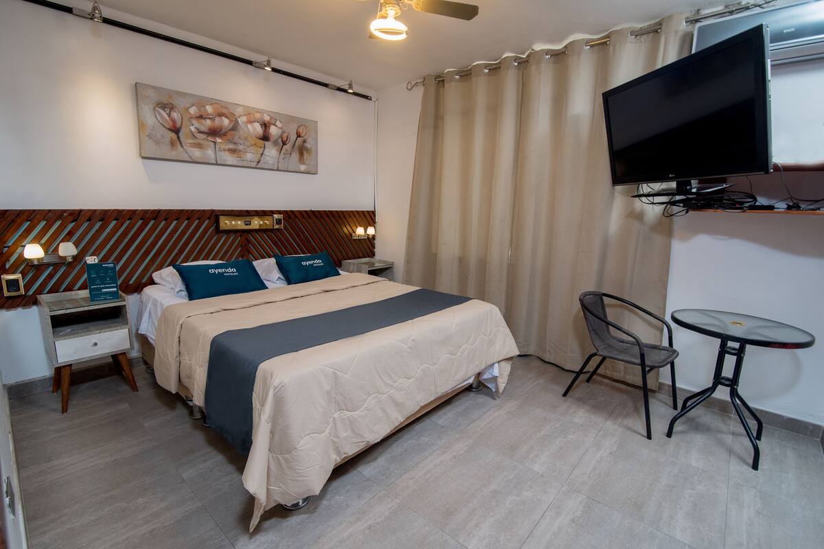 A hotel room with a bed, bedside tables, a desk, a chair, a TV, and a painting on the wall.