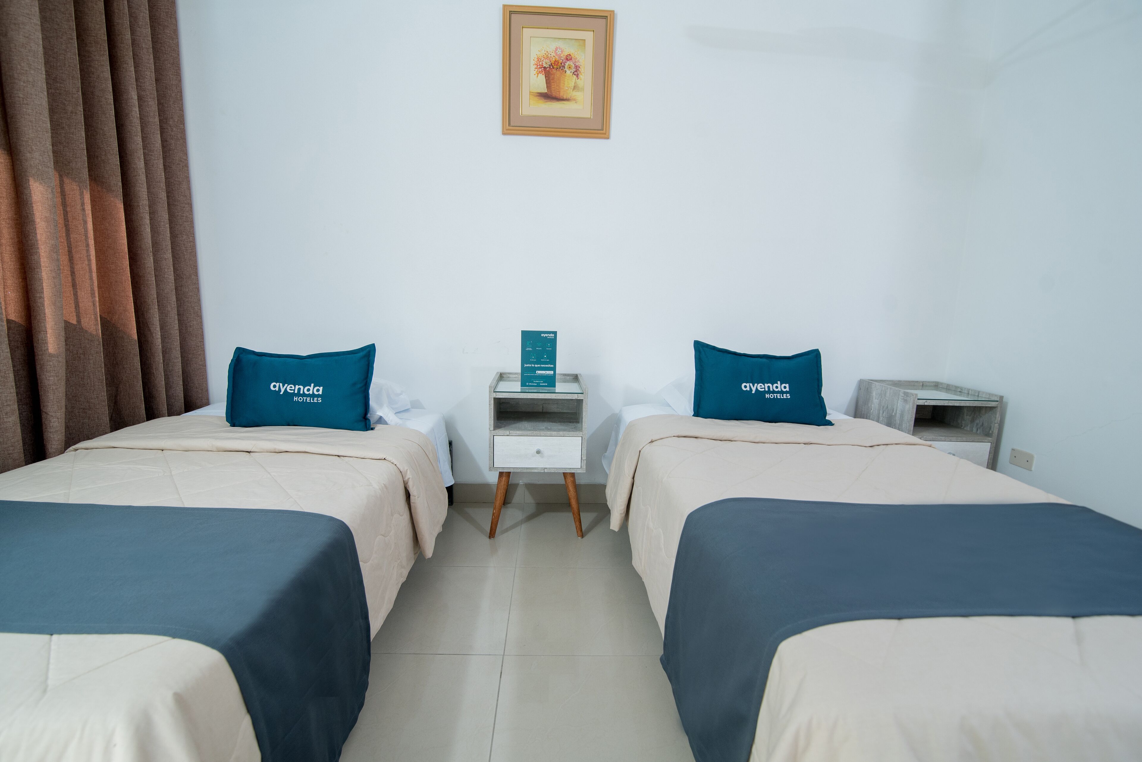 A hotel room with two beds, each with a blue pillow and a white nightstand.