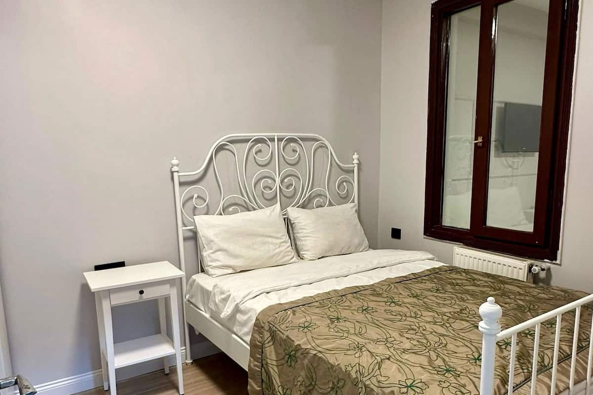 A bedroom with a white metal bed frame, a wooden bedside table, a beige floral bedspread, and a large window with a dark frame.