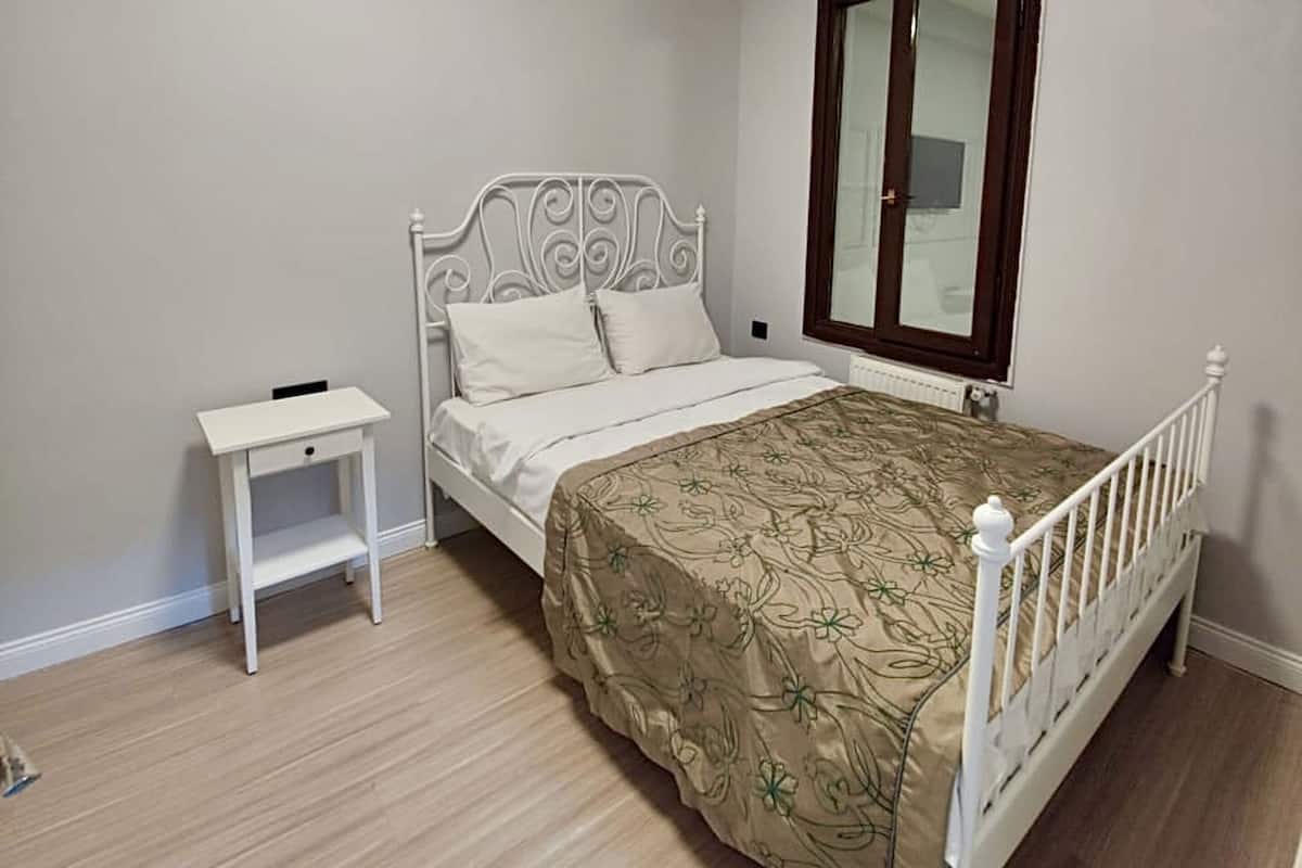 A bedroom with a single bed, a small bedside table, a window with a wooden frame, and a ceiling light.