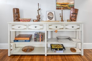 Una consola de madera blanca con varios cajones, una bandeja y elementos decorativos como un cuadro enmarco, una máscara de madera y una cesta.