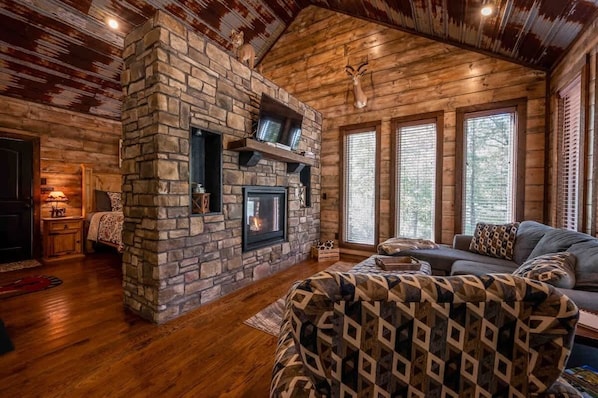 A cozy living room with a stone fireplace, wooden floors, and large windows.