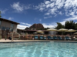 Une piscine avec des chaises longues et des parasols, un bâtiment avec une montagne en arrière-plan et un ciel dégagé.