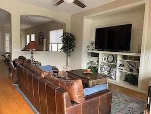 Un salon avec un long canapé en cuir marron, une table basse, un téléviseur posé sur une étagère blanche et une plante en pot.