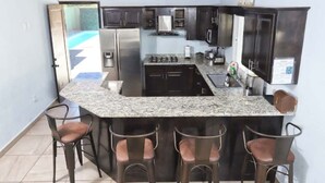A kitchen with granite countertops, dark cabinets, stainless steel appliances, and bar stools. - Relaxing Getaway w/ Pool, Billiards, Steps to the Beach – Perfect for Families! (Puerto Peñasco)