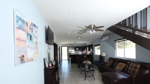 A spacious living area with a dark brown leather sofa, a coffee table, and a TV on the wall. There is a staircase leading to an upper level, and a kitchen area is visible in the background. - Relaxing Getaway w/ Pool, Billiards, Steps to the Beach – Perfect for Families! (Puerto Peñasco)