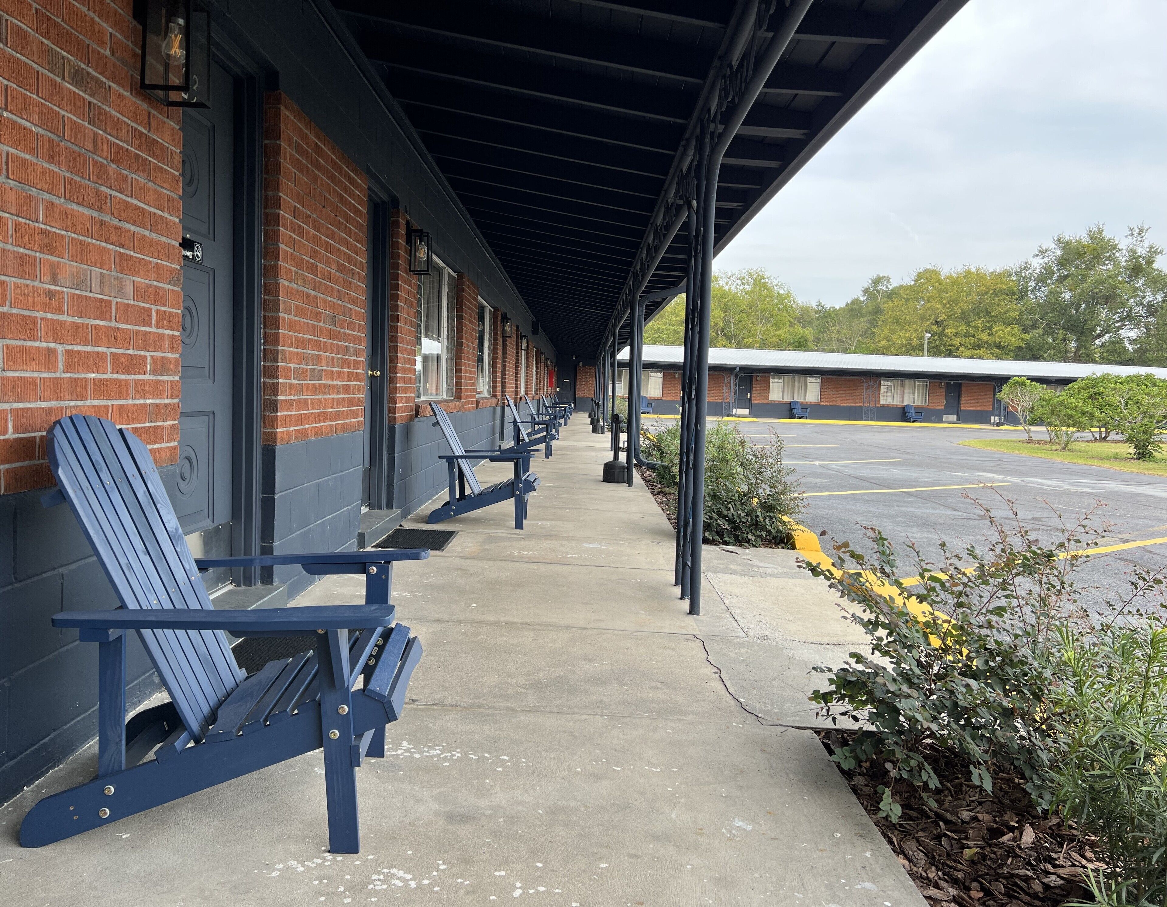 A covered outdoor seating area with blue Adirondack chairs, a brick building, and a parking lot.