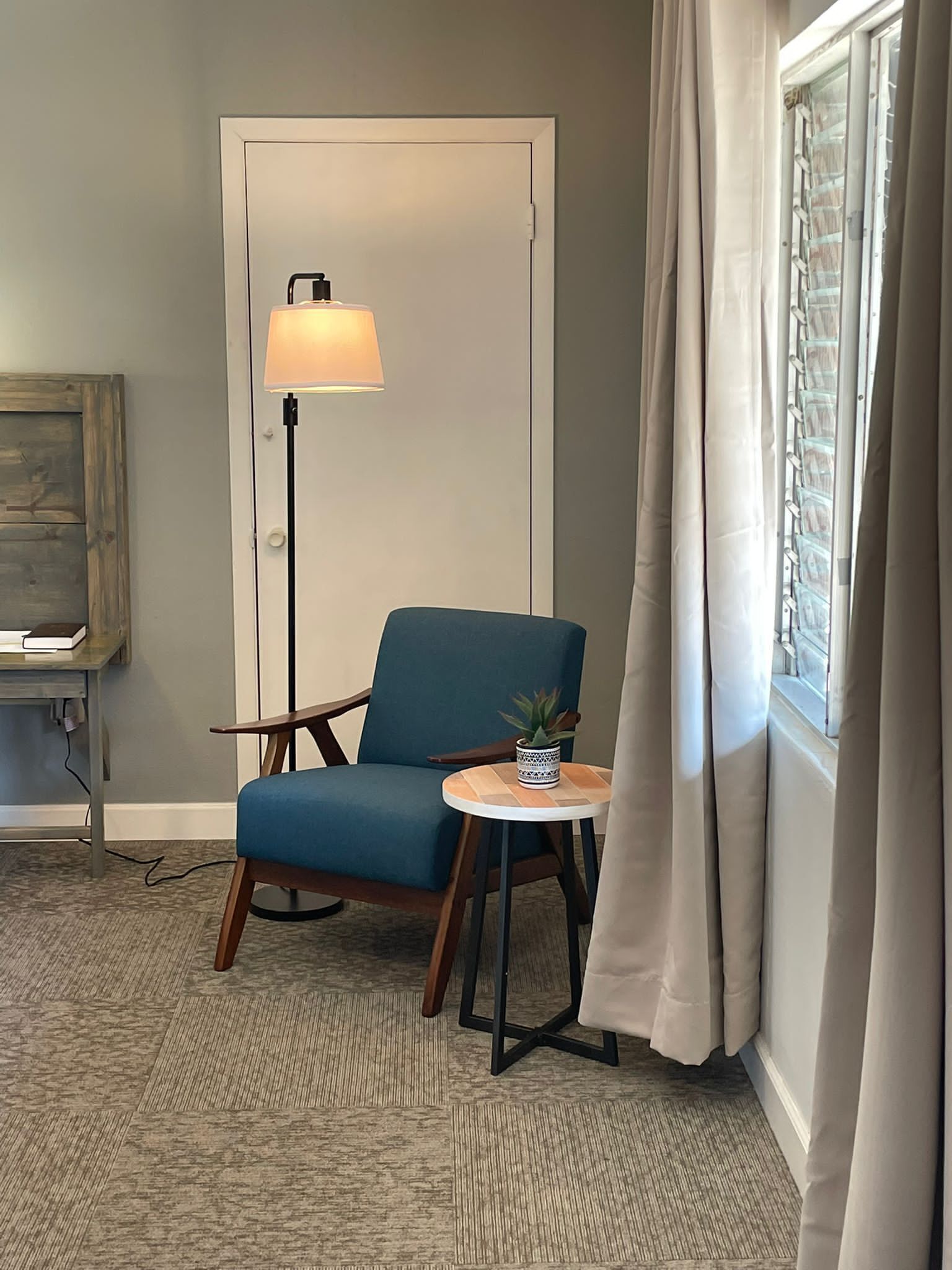 Ett rum med en blå fåt ölj, ett litet runt bord med en krukväxt, en golvlampa och en utsikt genom ett fönster med gardiner.