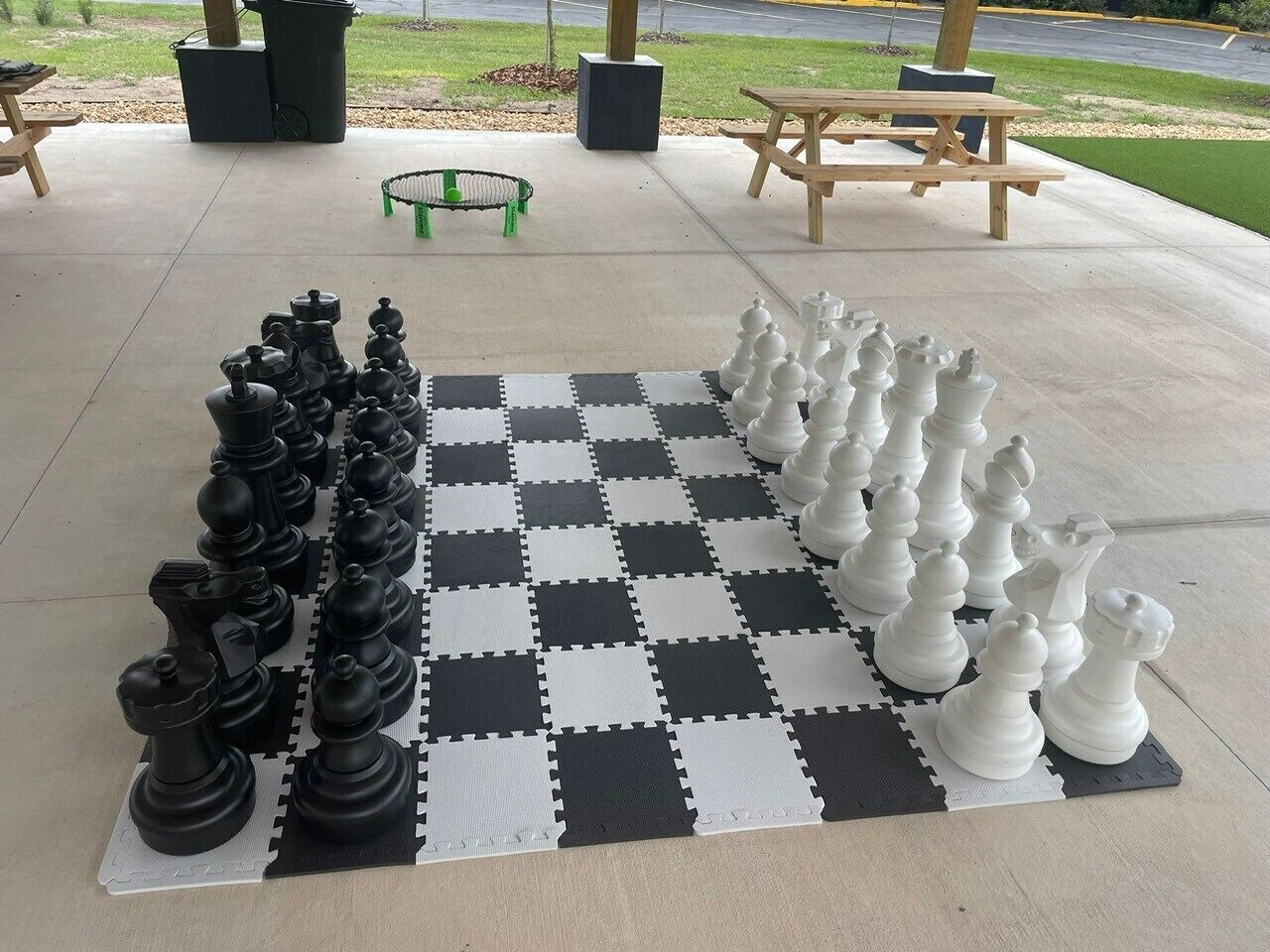 Een groot schaakbord buiten, opgesteld op een betonnen terras met een geruit kleed, omringd door tuinmeubels en een gazon op de achtergrond.