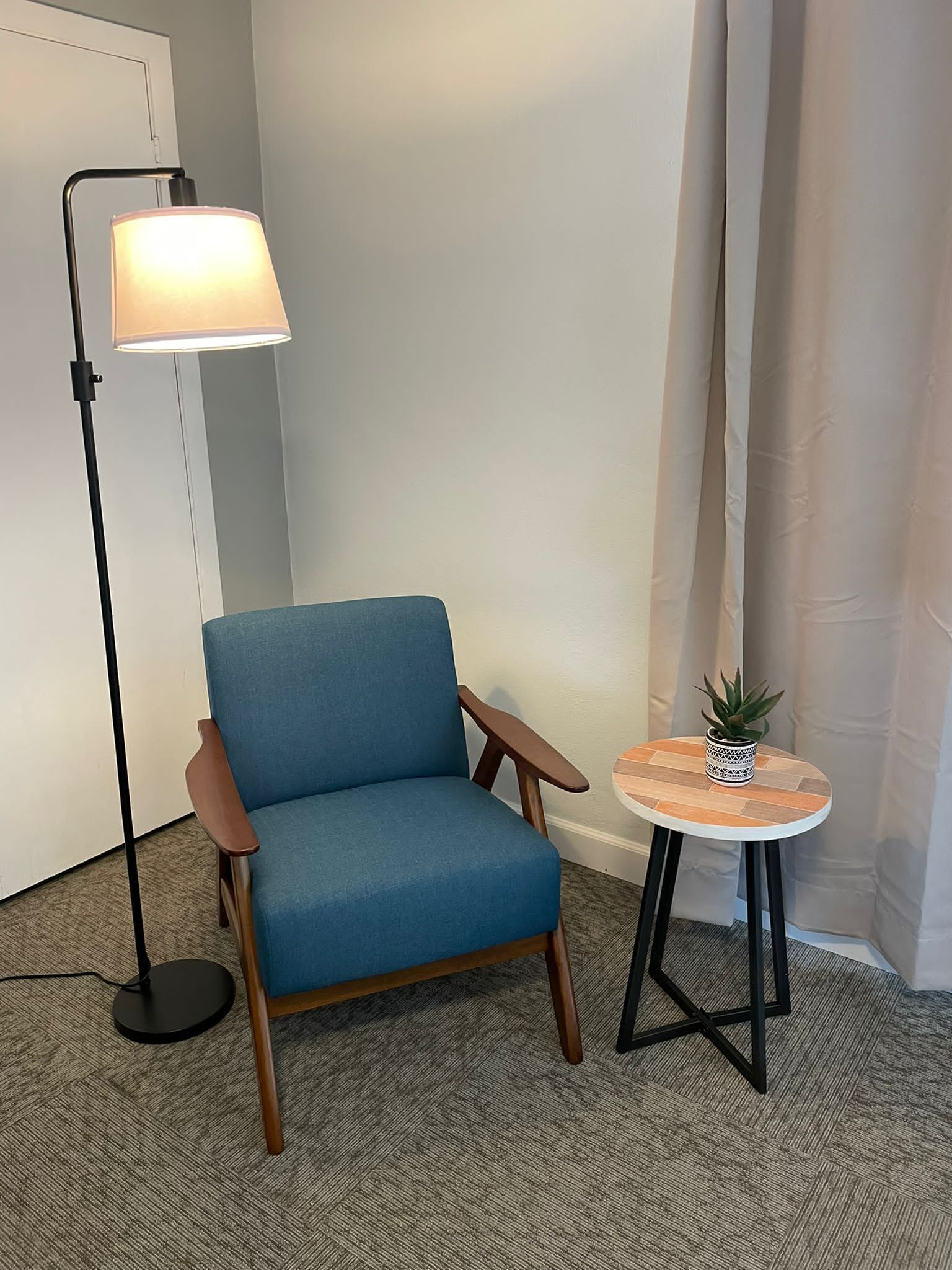 Una habitación con un sillón azul, una lámpara de pie, una pequeña mesa de madera con una planta y una cortina.