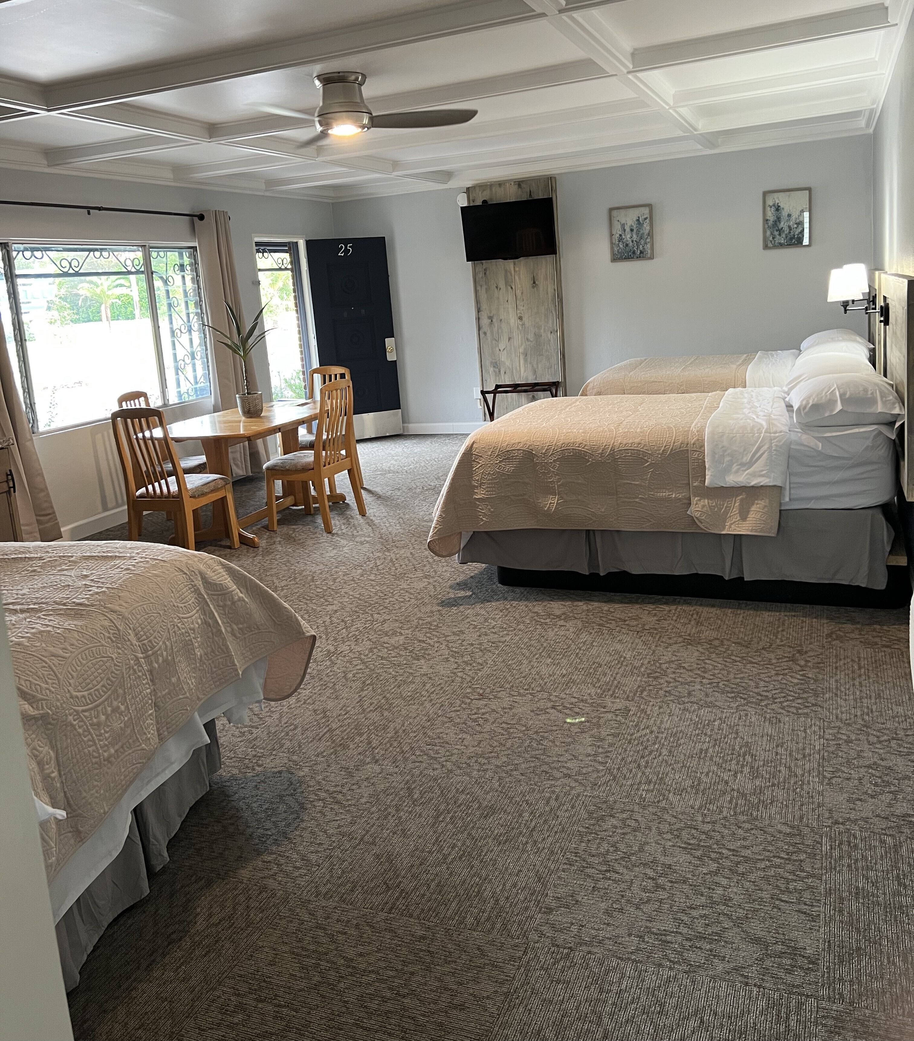 A hotel room with two beds, a dining table with chairs, a ceiling fan, and a view of the outdoors through a window.