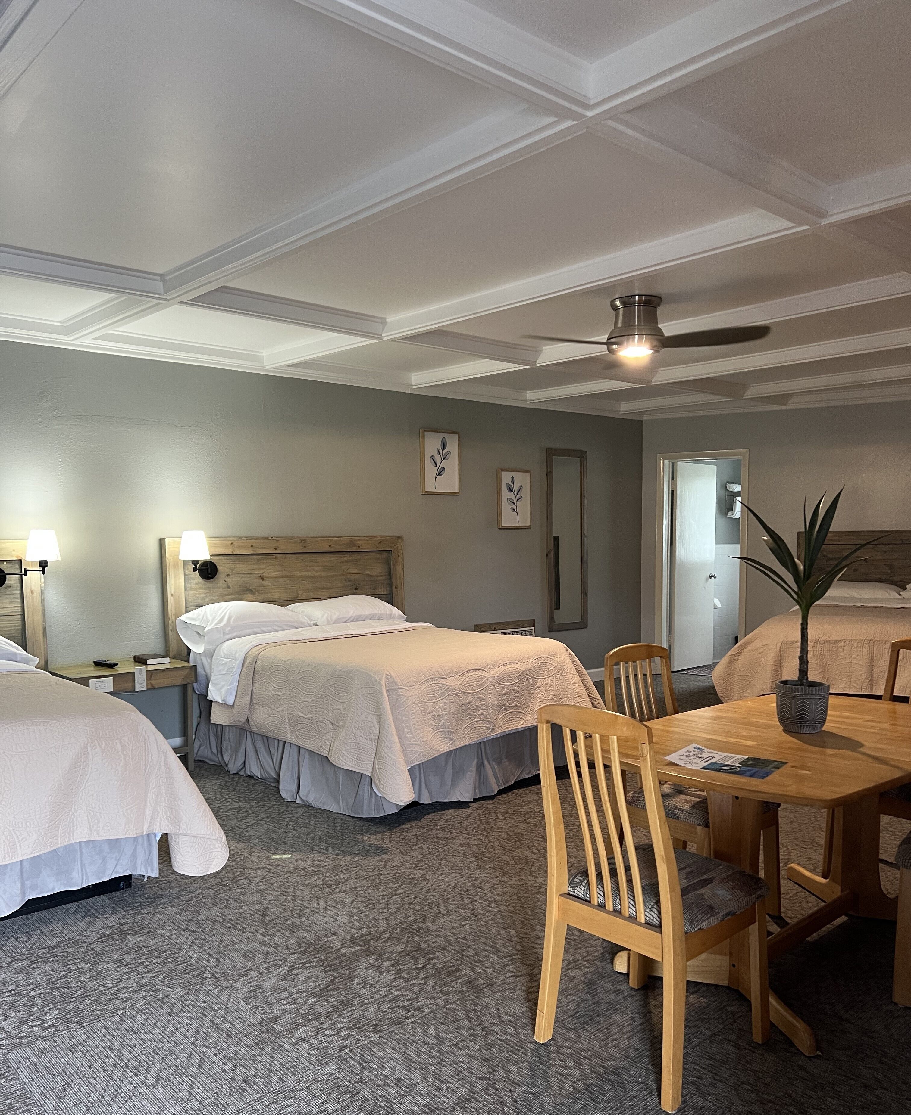 A hotel room with two beds, a dining table with chairs, a ceiling fan, and a potted plant.
