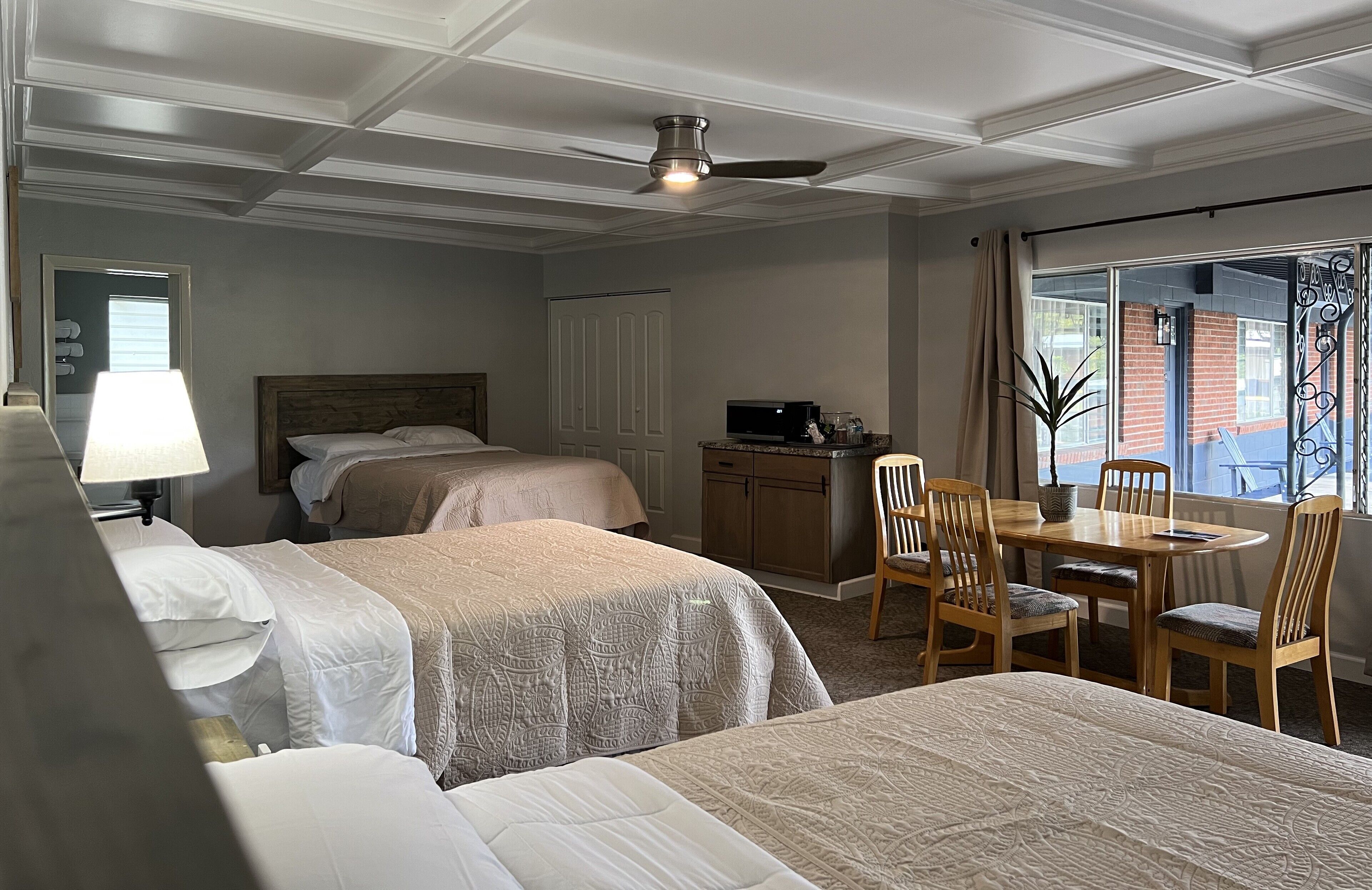 A hotel room with two beds, a dining table with chairs, a ceiling fan, and a view of the outside.