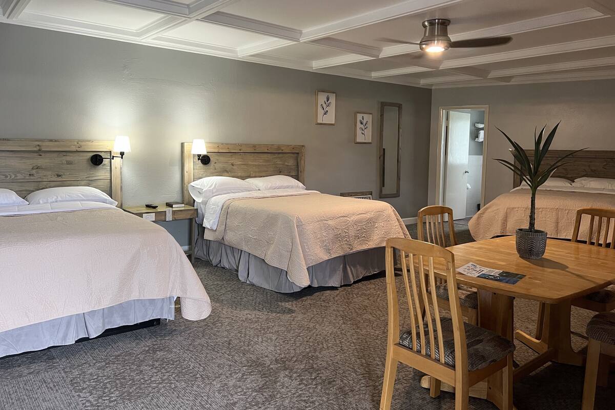 A hotel room with three single beds, a wooden headboard, a ceiling fan, and a dining table with chairs.