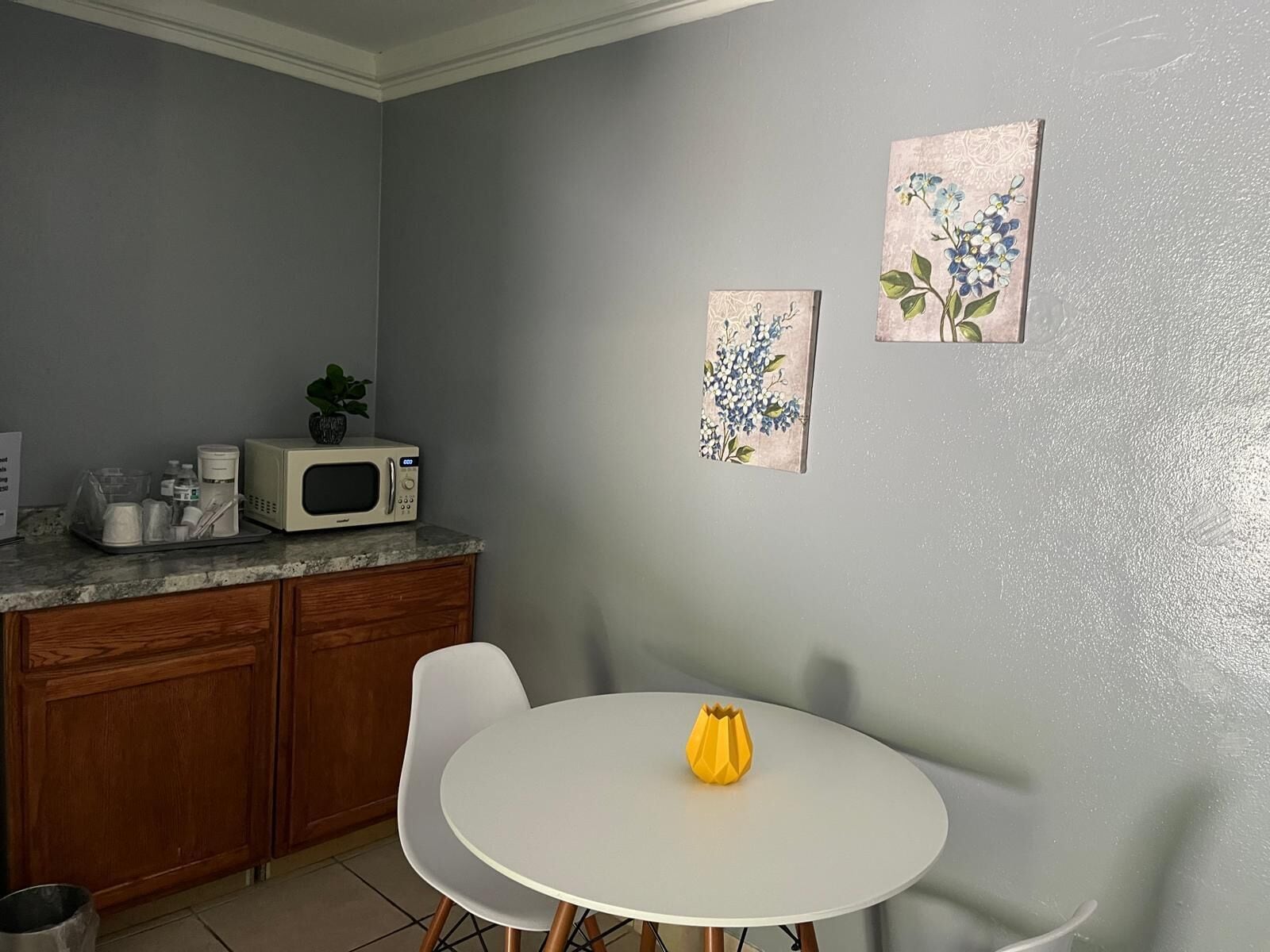 A kitchen with a microwave, coffee maker, and a small potted plant on the counter. There is a white table with a yellow vase and two framed floral artworks on the wall.