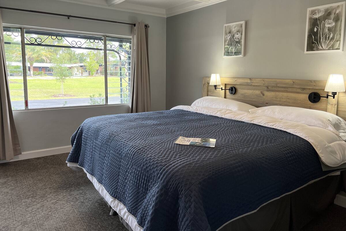 A neatly made bed with a blue quilt, two bedside lamps, and a window with curtains.