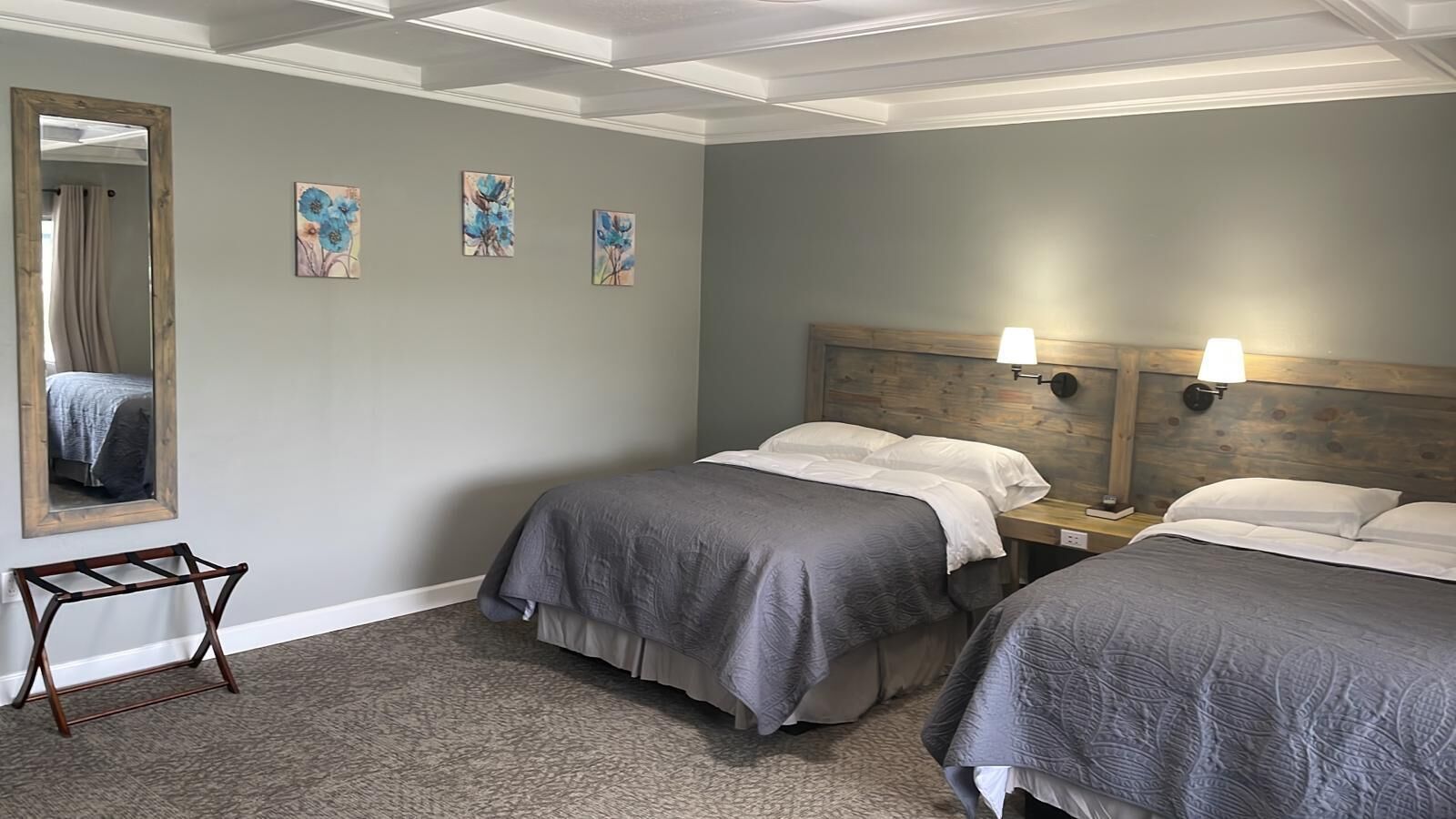 Habitación de hotel con dos camas, cabecera de madera, mesita de noche, espejo y tres cuadros florales en la pared.