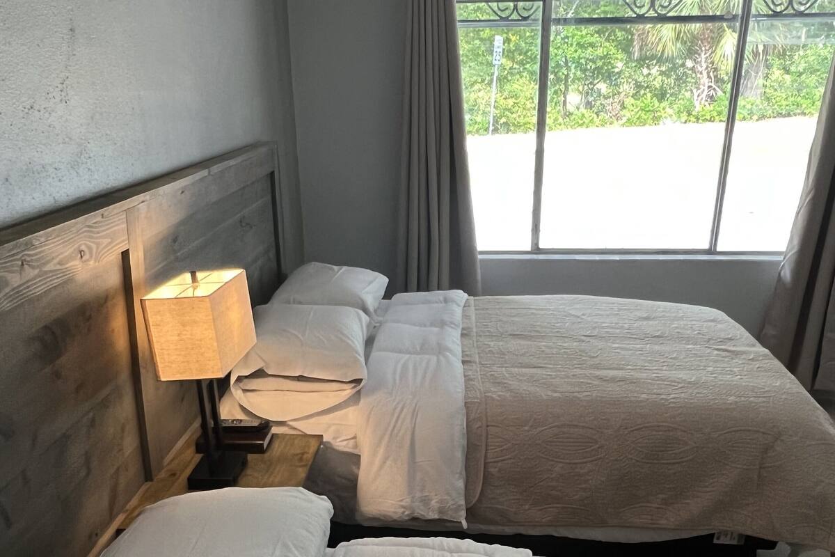 A hotel room with two beds, a nightstand with a lamp, and a window with a view of greenery.