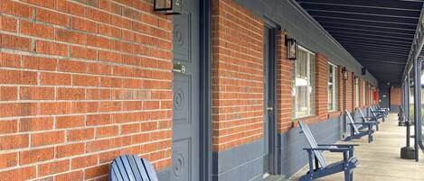 A row of blue Adirondack chairs along a covered walkway with brick walls and doors.