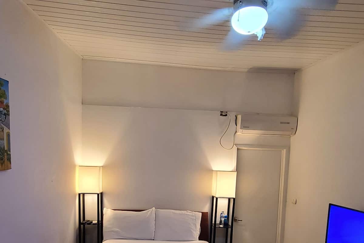 A hotel room with a bed, two bedside lamps, a ceiling fan, an air conditioning unit, a TV, and a water bottle on a nightstand.