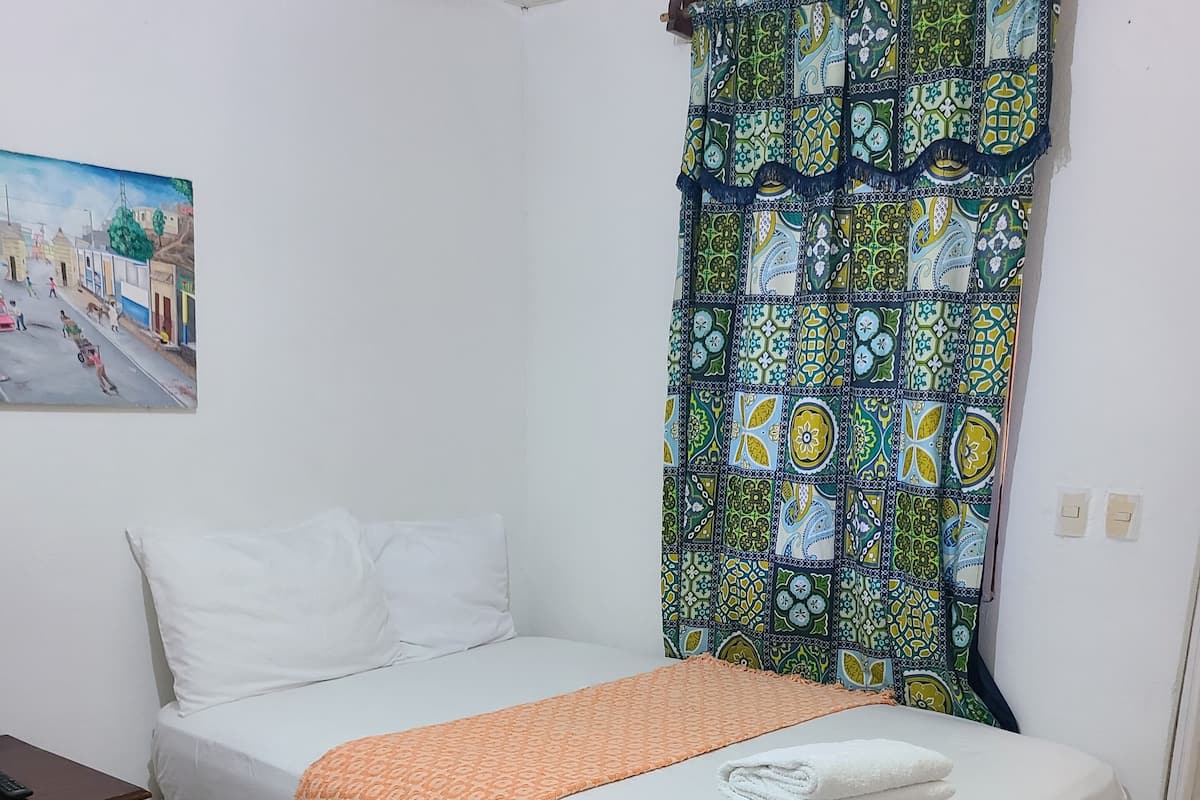 A bedroom with a bed, a nightstand, a painting on the wall, and a patterned curtain.