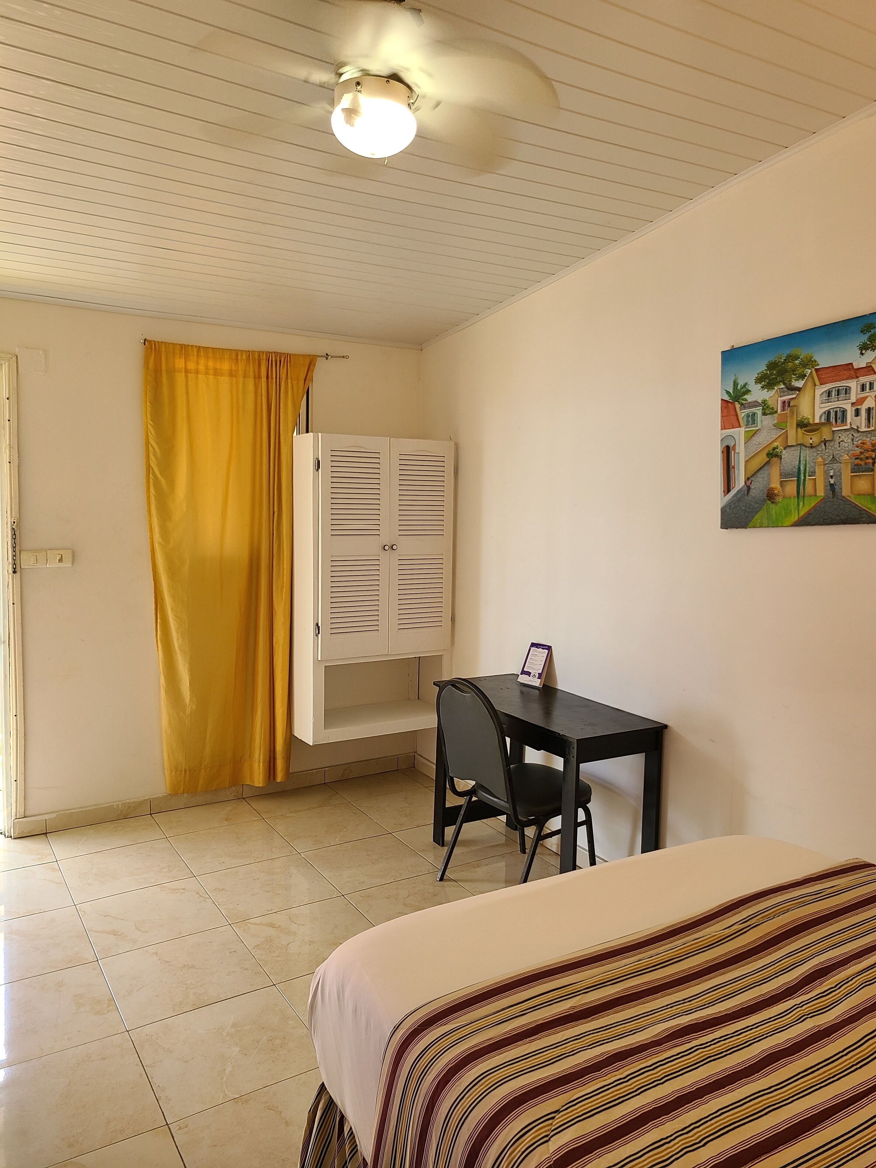 A room with a bed, a desk, a chair, a ceiling fan, a yellow curtain, and a painting on the wall.