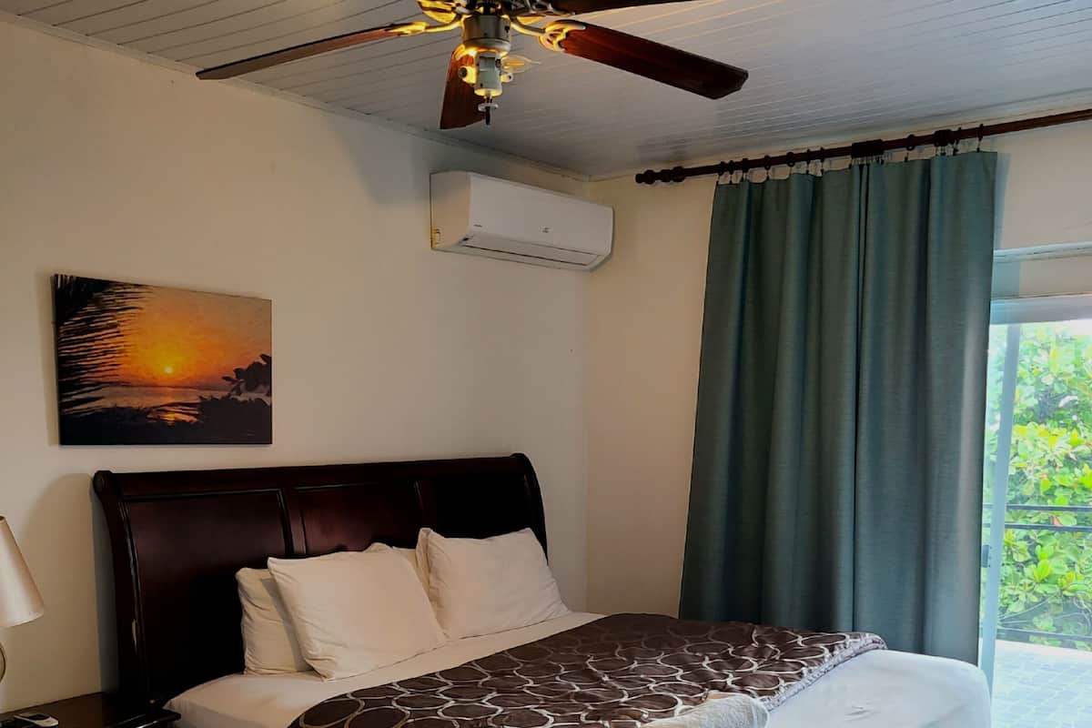 A bedroom with a bed, a ceiling fan, an air conditioning unit, a nightstand, a lamp, a painting of a sunset, and a view through a window with curtains.
