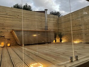 Une terrasse en bois dotée d’un sauna intégré, de plantes en pot et d’un appareil de chauffage pour sauna en métal.