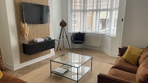 A modern living room with a flat-screen TV mounted on a wooden panel wall, a tripod lamp, a black console table with decorative items, a glass coffee table, brown sofas, and a large window with blinds. - Luxury hotel style 2-bed home with hot tub central location (pet friendly) (Wooler)