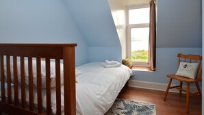 Een slaapkamer met een schuine plafond, een bed met witte lakens, een houten stoel met een kussen en een raam met gordijnen.