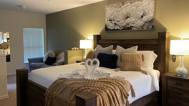 A neatly made bed with a swan-shaped pillow arrangement, a large floral artwork above the bed, and a bedside lamp on each side. - Wish Upon A Star 5 bed pool/jacuzzi spa heat included (Clermont)