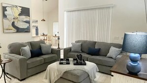 A modern living room with two grey sofas, a white ottoman, a side table with a lamp, and a painting on the wall. - Wish Upon A Star 5 bed pool/jacuzzi spa heat included (Clermont)