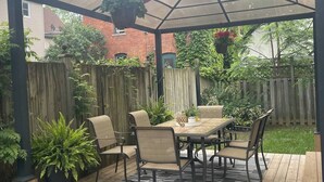 Un patio cubierto con una mesa y sillas de madera, rodeado de plantas en macetas y una cerca de madera.