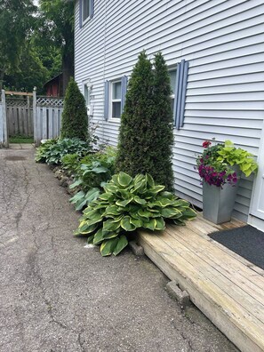 A house with a gravel driveway, a wooden deck, and a row of plants including hostas and a tall conifer. - Spacious Niagara Escape 3 BR Private Backyard (Niagara Falls)