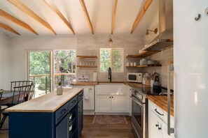 Une cuisine moderne avec des poutres en bois, des armoires blanches, un îlot central et une vue sur la verdure à l’extérieur.