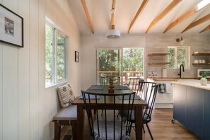 Un espace repas comprenant une table et des chaises en bois, un banc et une vue sur l’extérieur grâce à de grandes fenêtres.