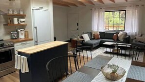 A modern kitchen with a central island, a dining area with a table and chairs, a sofa, and a large window with curtains. - All New, 3 Bdrms, HOT TUB, near town and trails (Idyllwild-Pine Cove)