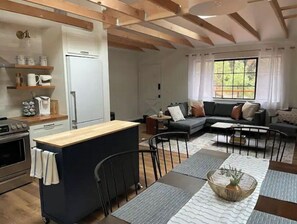 A modern kitchen with a central island, a dining area with a table and chairs, a sofa, and a large window with curtains.