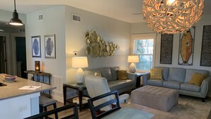A modern living room with a grey sofa, a coffee table, and a dining area with chairs. There is a decorative wall piece and a unique hanging light fixture. - Funky coconut luxury apartment (Dunedin)